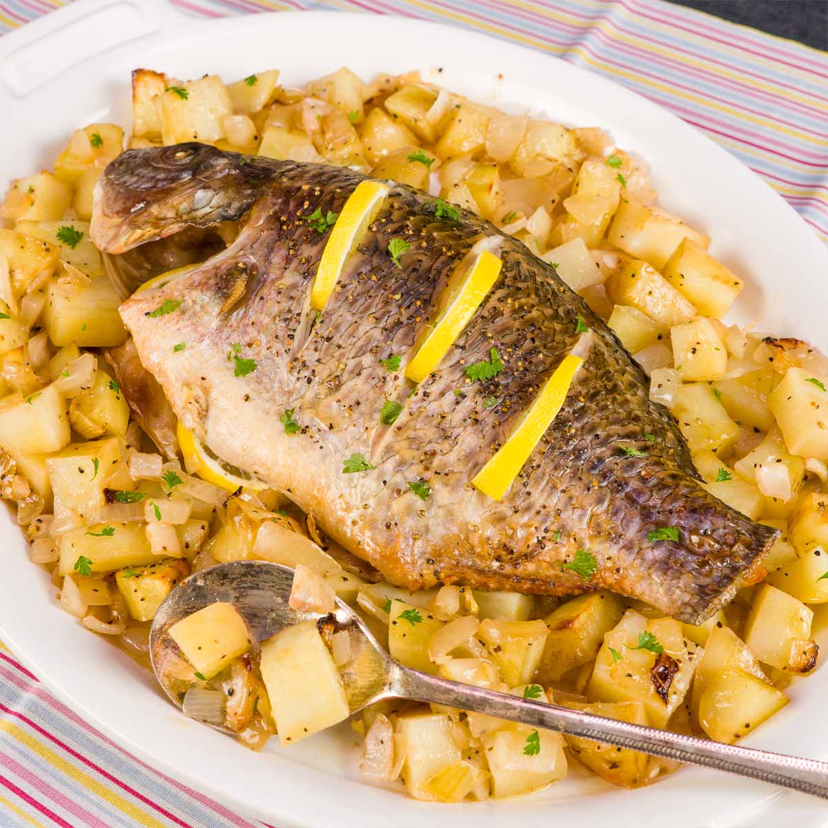 Baked whole tilapia with lemon slices on top, served on a white platter over roasted potatoes and onions.