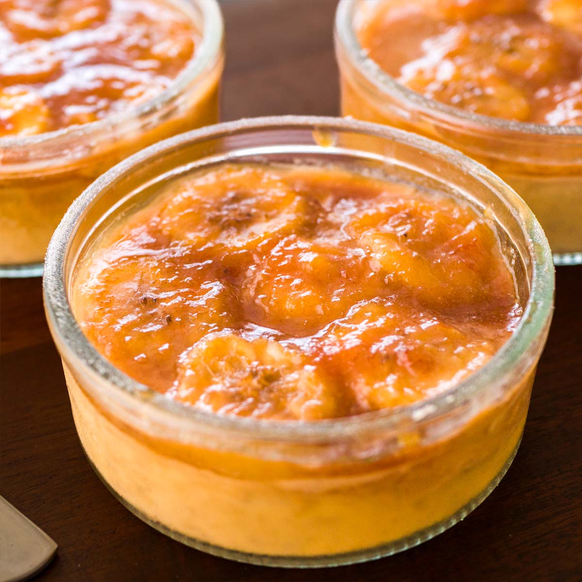 Banana Foster pudding in small glass bowls, topped with glossy caramelized banana sauce on a wooden surface.