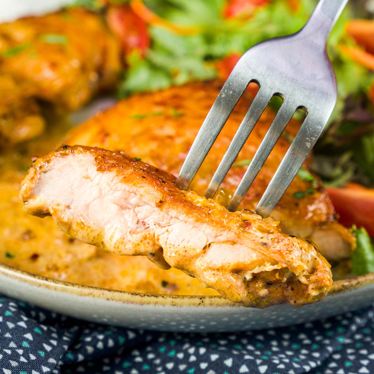 Fork holding a bite of tender chicken thigh coated in creamy sauce, with the plated chicken and salad blurred in the background. 