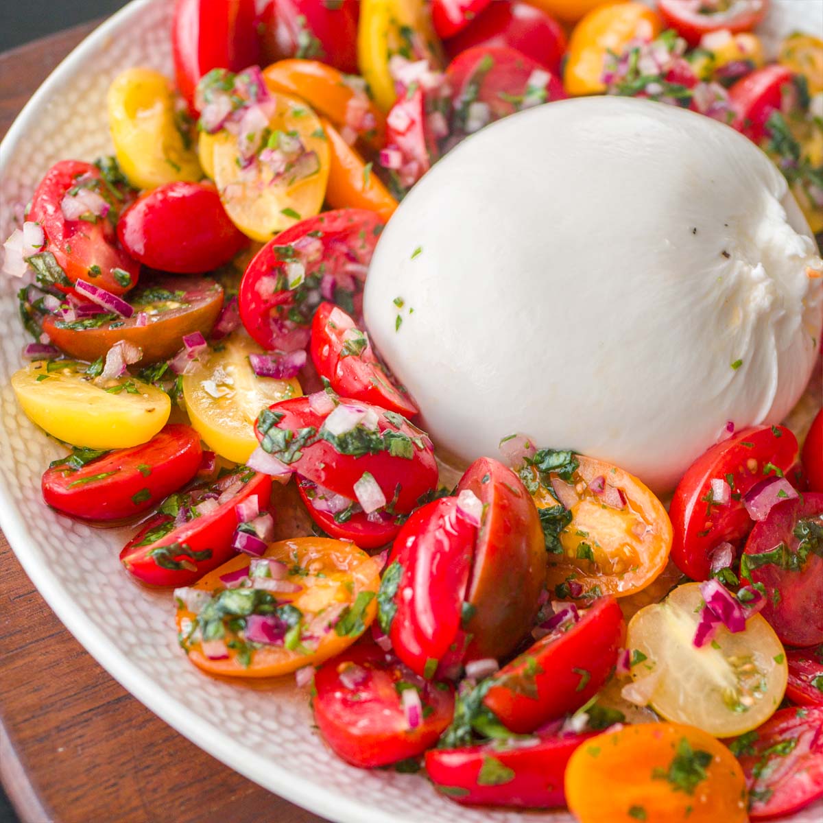 Close-up of Tomato Burrata Salad with colorful cherry tomatoes tossed in herbs and red onion, surrounding a fresh ball of burrata on a white plate.