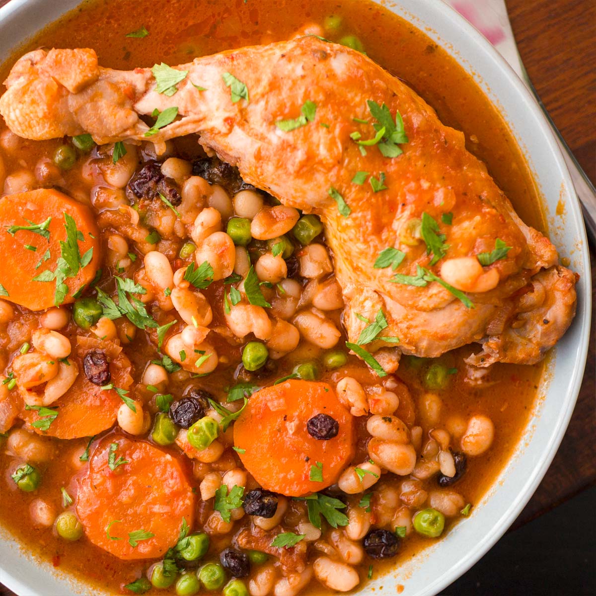 Chicken stew with white beans, carrots, peas, and raisins in a rich tomato sauce, served with fresh parsley on top.