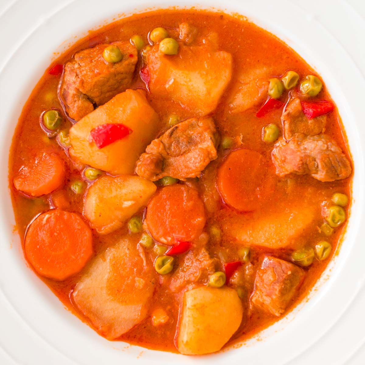 Spanish Pork and Potato Stew with tender chunks of pork, potatoes, carrots, peas, and red peppers simmered in a rich tomato sauce, served in a white bowl. 