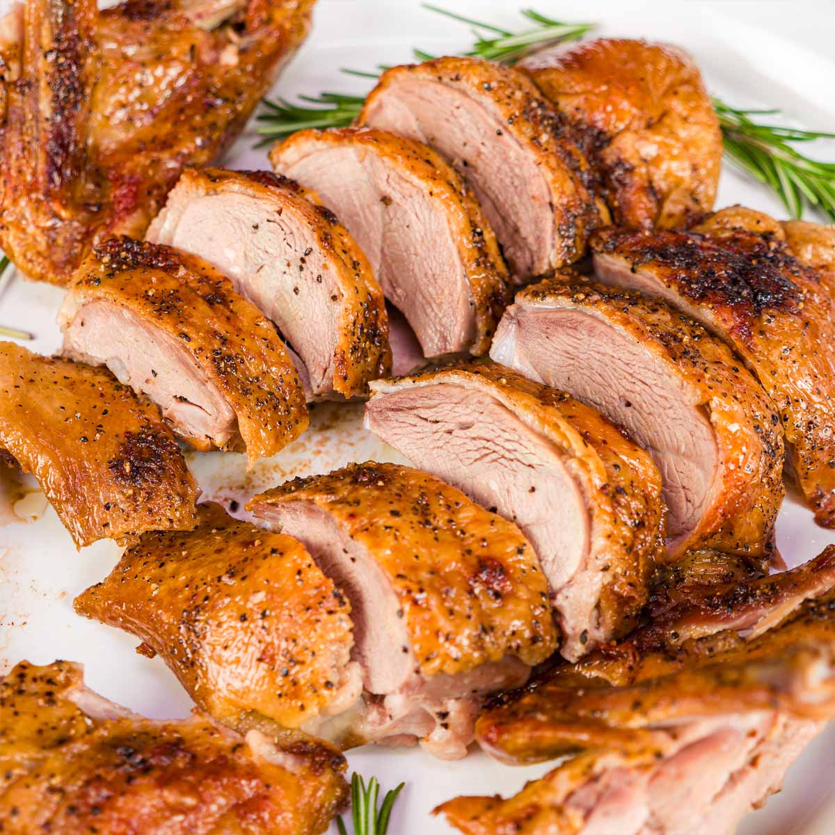 Close-up of roasted duck breast sliced into thick pieces, showing juicy meat and crisp, golden skin, served on a platter with rosemary.
