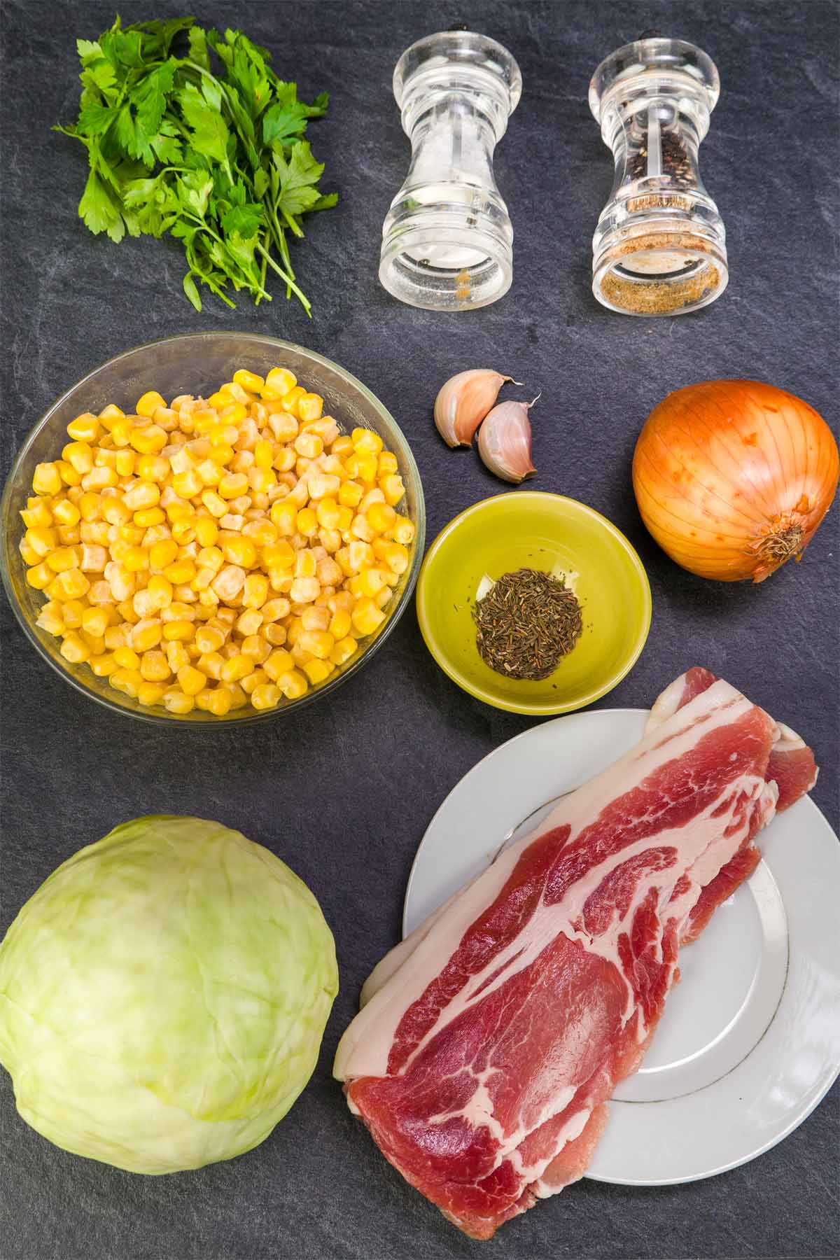 Ingredients for fried cabbage with corn and bacon: cabbage, corn kernels, bacon strips, onion, garlic, oil, salt, pepper, and fresh herbs on a dark surface. 