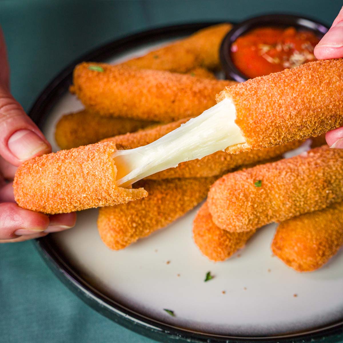 A close-up of two hands pulling apart a crispy homemade mozzarella stick, revealing the gooey melted cheese inside, with more mozzarella sticks on the plate. 