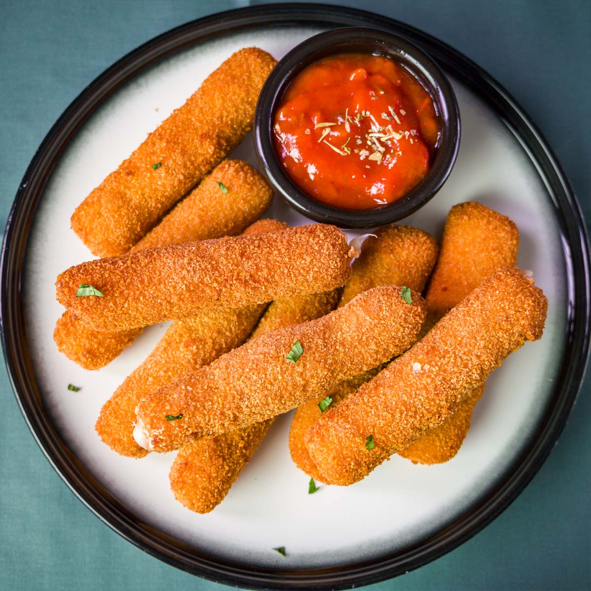 A plate of golden, crispy homemade mozzarella sticks served with a small bowl of marinara sauce for dipping.