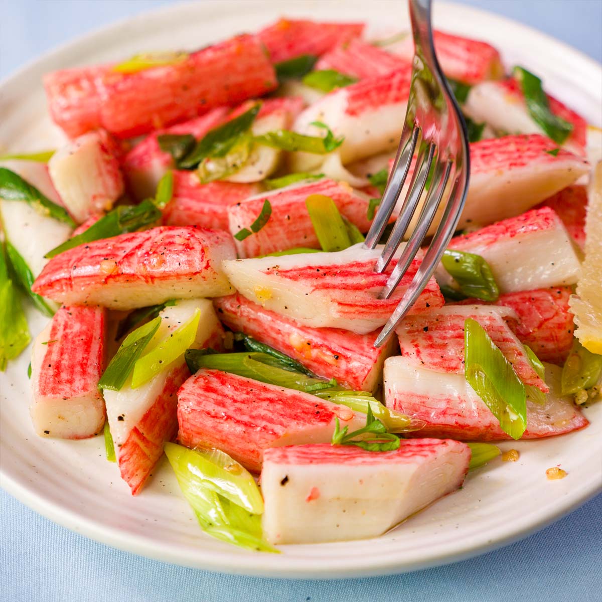 Fork lifting a piece of sautéed imitation crabmeat tossed in garlic butter with green onions and black pepper, served on a white plate with a lemon wedge. 