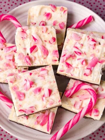 Homemade white chocolate peppermint bark squares topped with crushed candy canes on a plate with pink peppermint sticks.