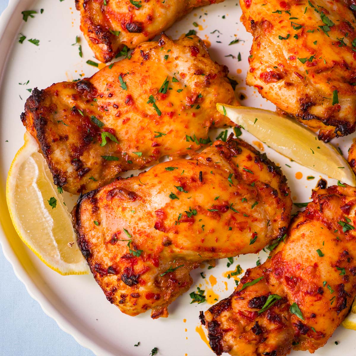 Close-up of baked harissa chicken thighs with caramelized edges, garnished with parsley and served with lemon wedges.