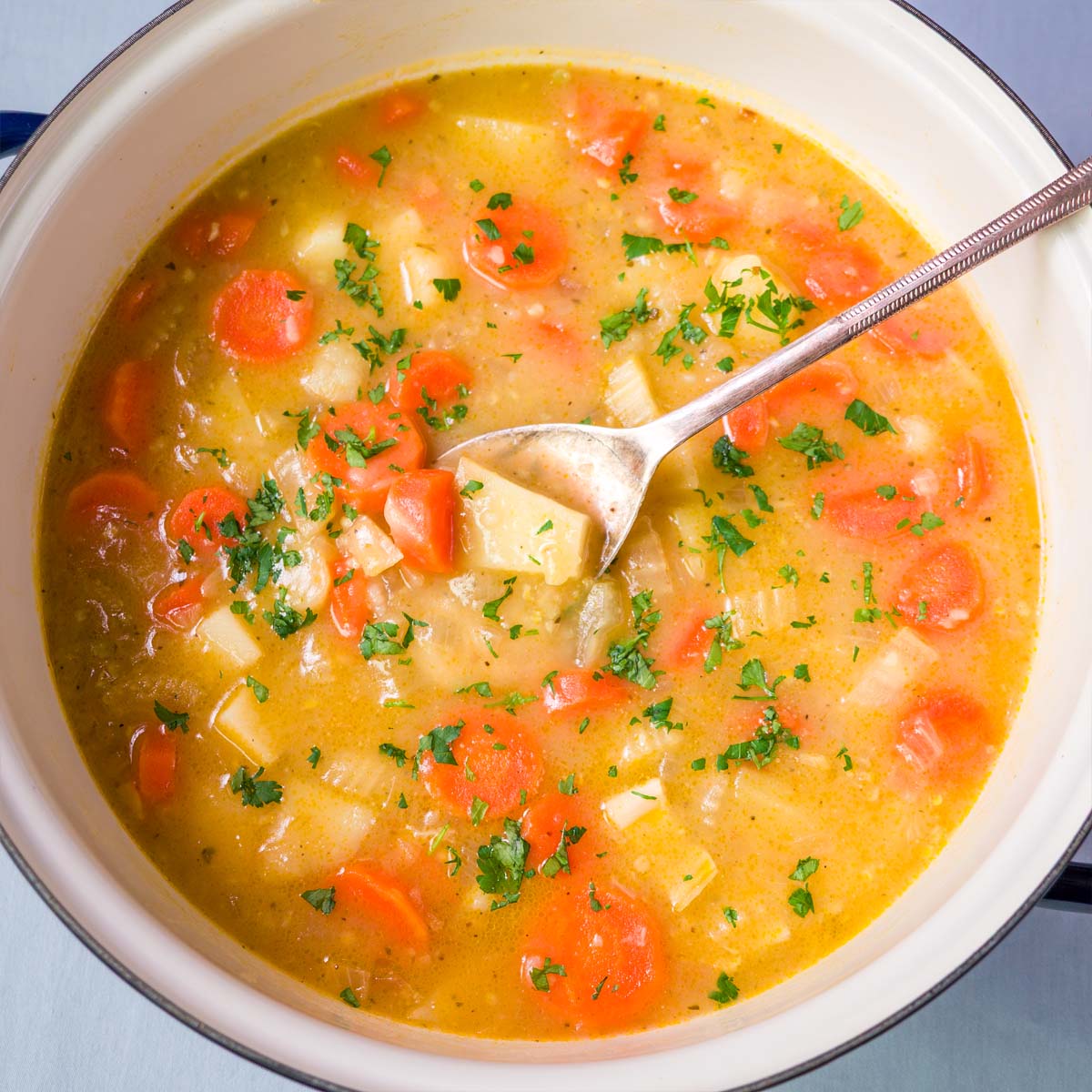 Close-up of easy carrot potato soup with soft carrots, diced potatoes, celery, and chopped parsley in a warm golden broth, with a spoon in the pot.