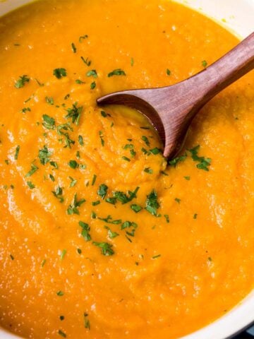 Creamy carrot parsnip soup in a bowl topped with chopped parsley, with a wooden spoon scooping the soup