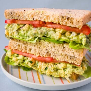 Chickpea salad sandwich with Greek yogurt on whole grain bread, layered with lettuce and sliced tomato on a plate.