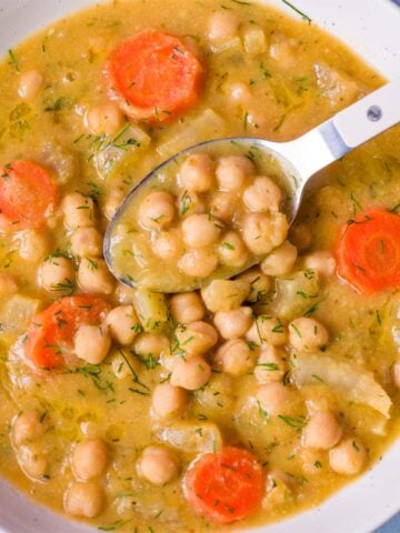 Spoon scooping Greek chickpea soup (Revithia) with chickpeas and carrot slices from a white bowl in a golden herb-speckled broth.