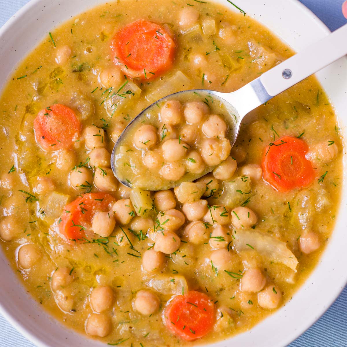 Spoon scooping Greek chickpea soup (Revithia) with chickpeas and carrot slices from a white bowl in a golden herb-speckled broth.