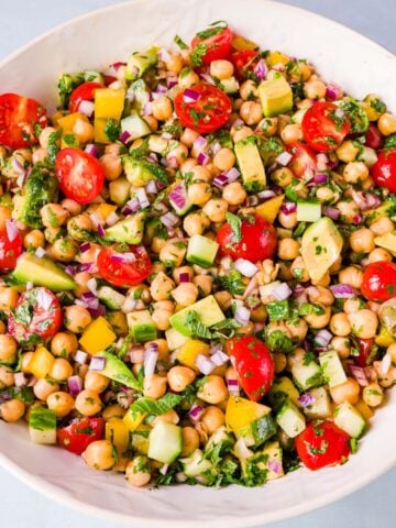 Mediterranean chickpea salad with avocado, cherry tomatoes, cucumber, red onion, herbs, and bell pepper in a white bowl.