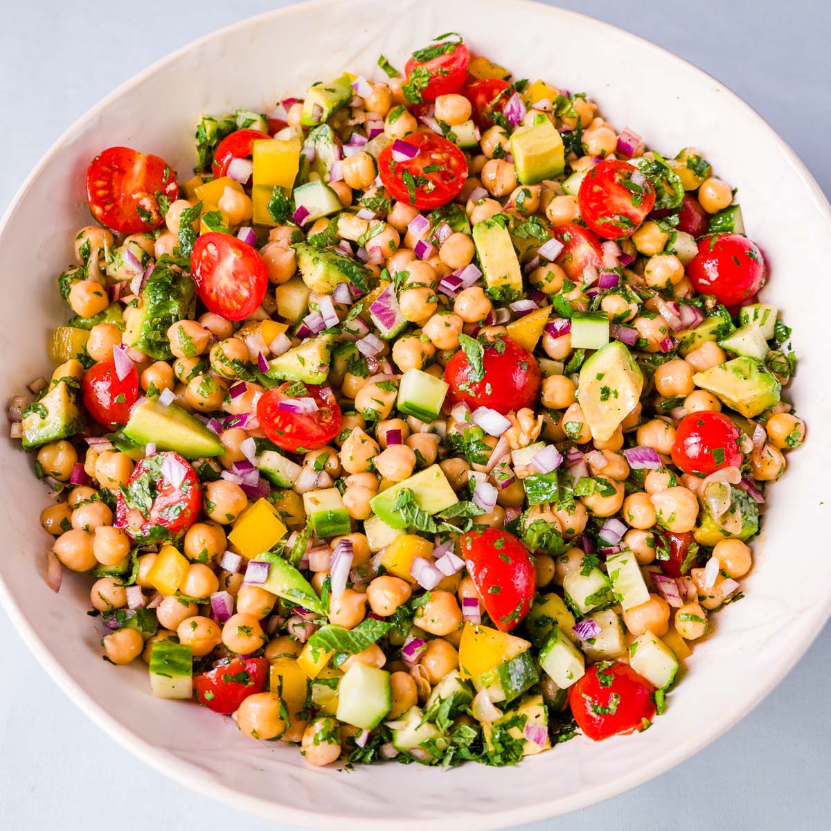 Mediterranean chickpea salad with avocado, cherry tomatoes, cucumber, red onion, herbs, and bell pepper in a white bowl.