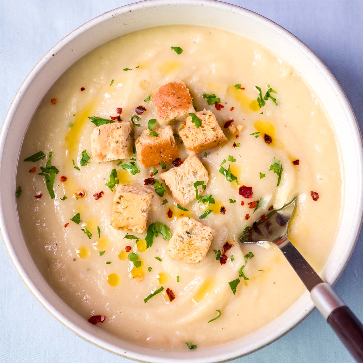 Bowl of creamy parsnip soup garnished with croutons, chopped parsley, red pepper flakes, and olive oil, with a spoon resting on the side