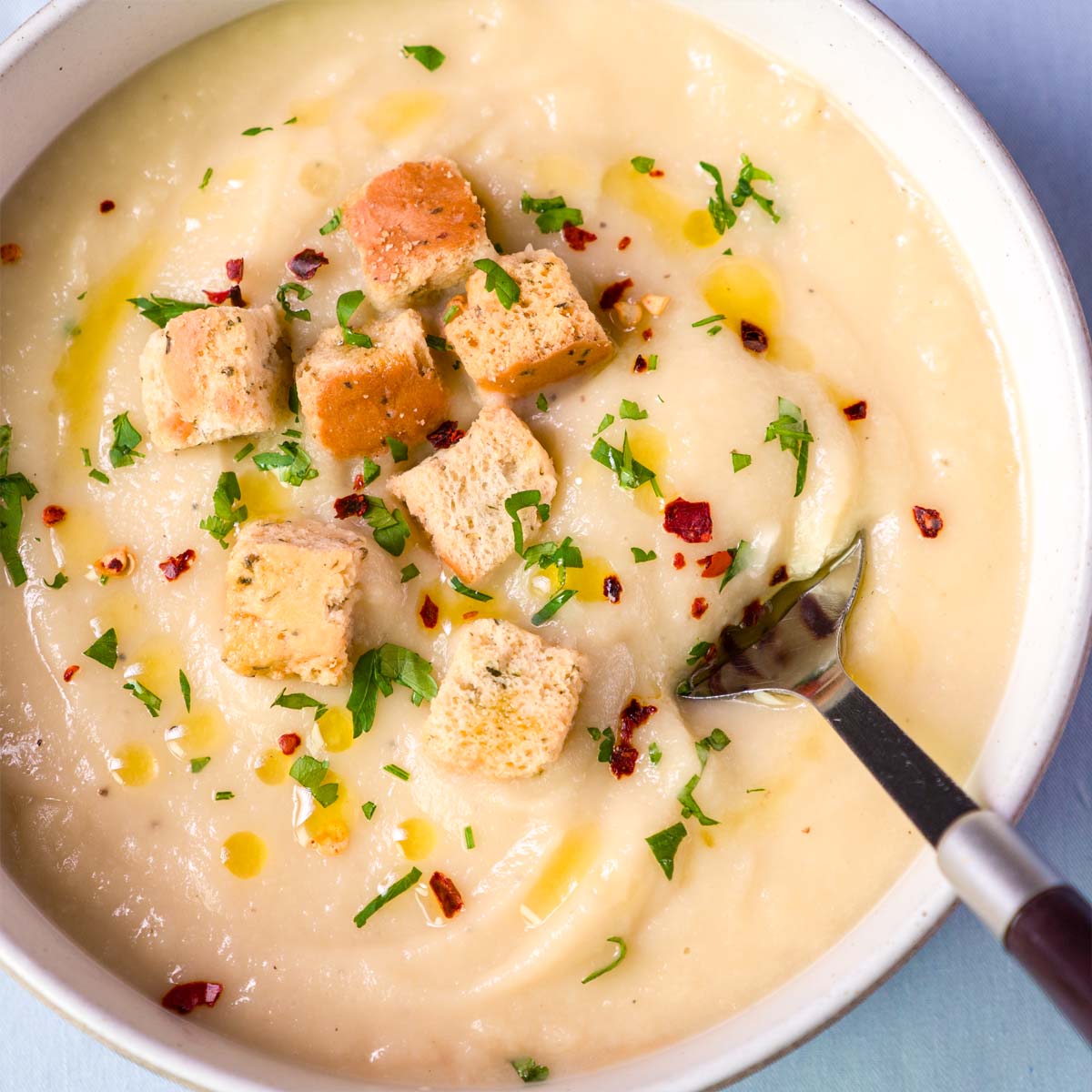 Creamy parsnip soup topped with croutons, chopped parsley, red pepper flakes, and a drizzle of olive oil, with a spoon in the bowl
