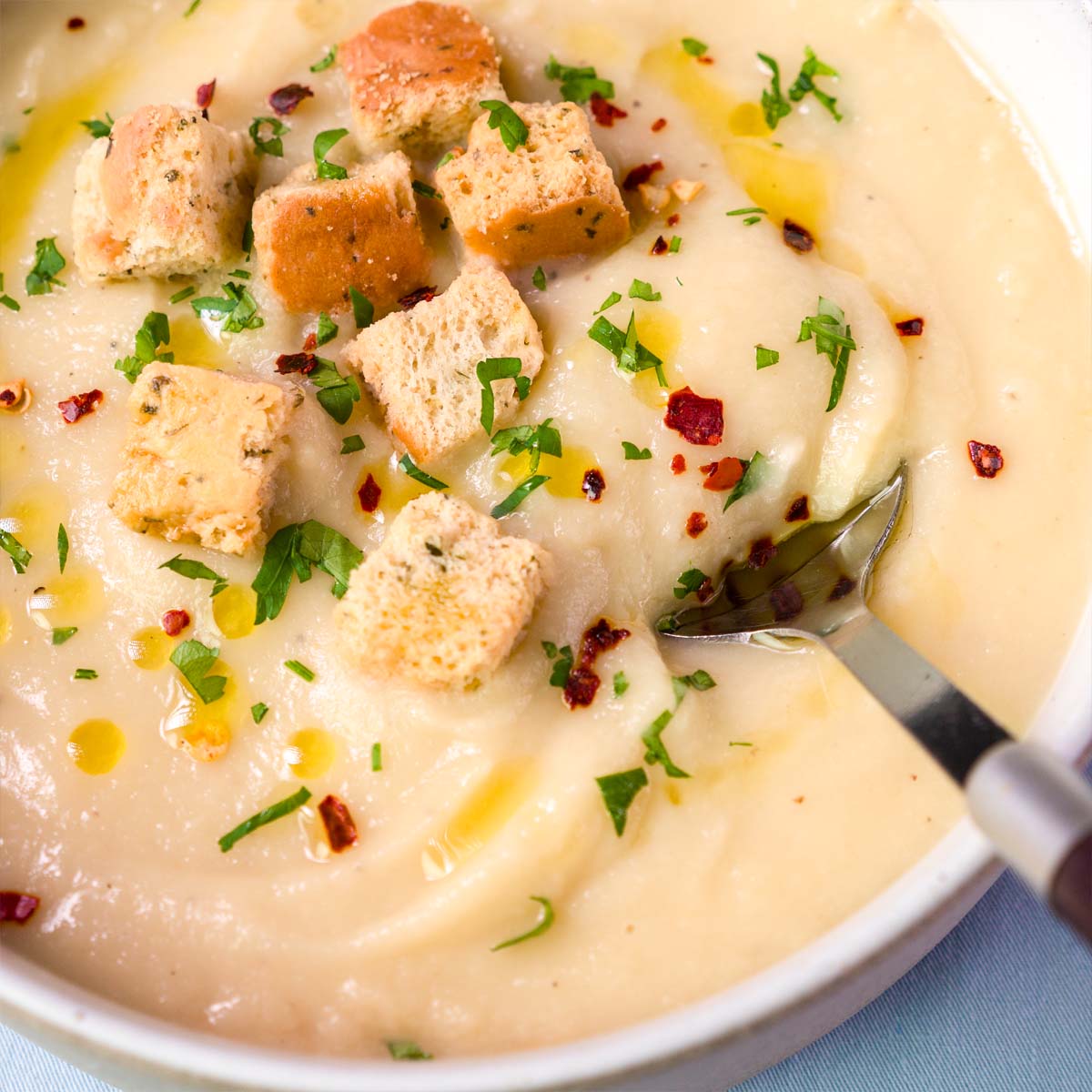 Close-up of creamy parsnip soup topped with toasted croutons, chopped parsley, red pepper flakes, and a drizzle of olive oil, with a spoon dipping in