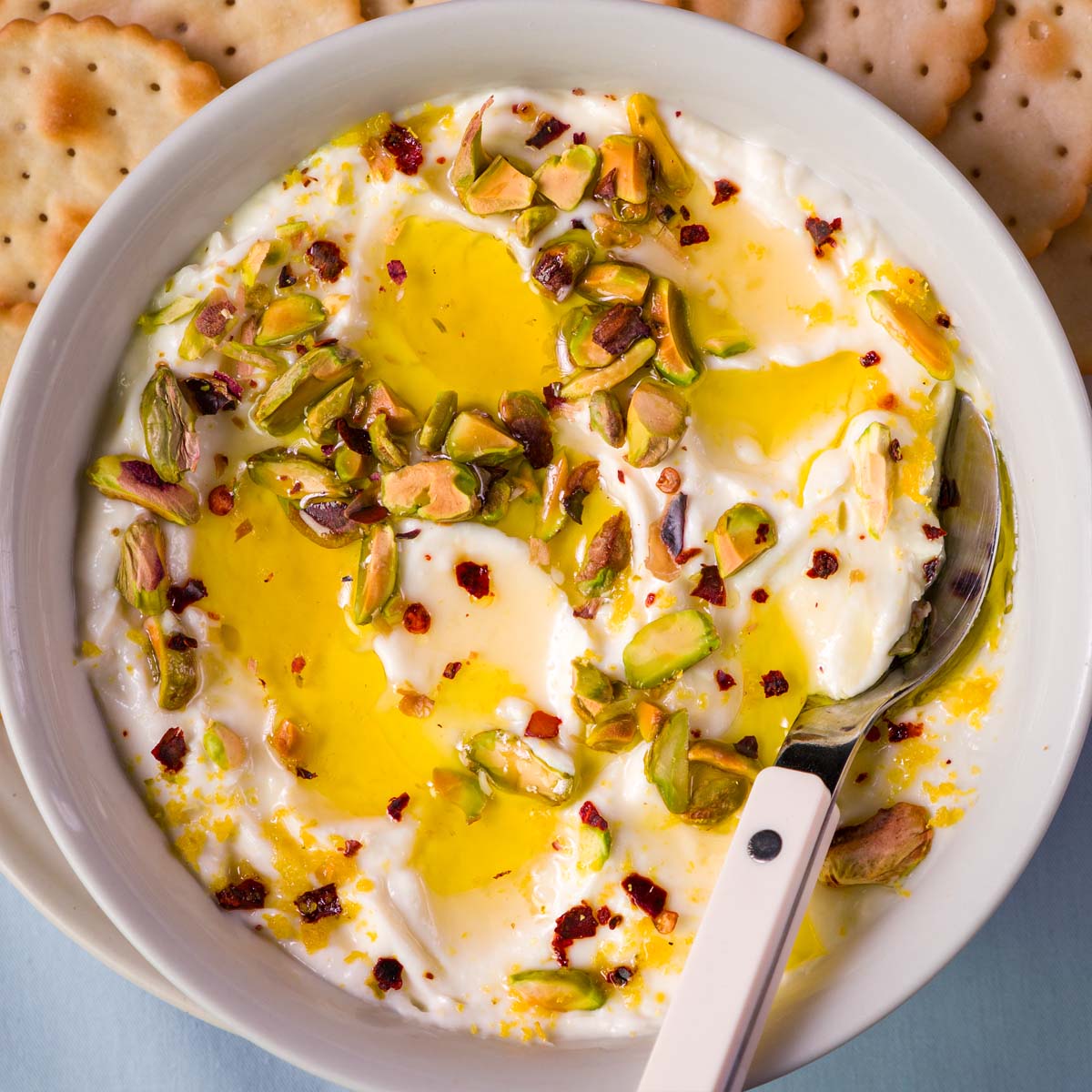 Close-up of whipped ricotta dip with olive oil, honey drizzle, chopped pistachios, and red pepper flakes, served in a white bowl with a spoon