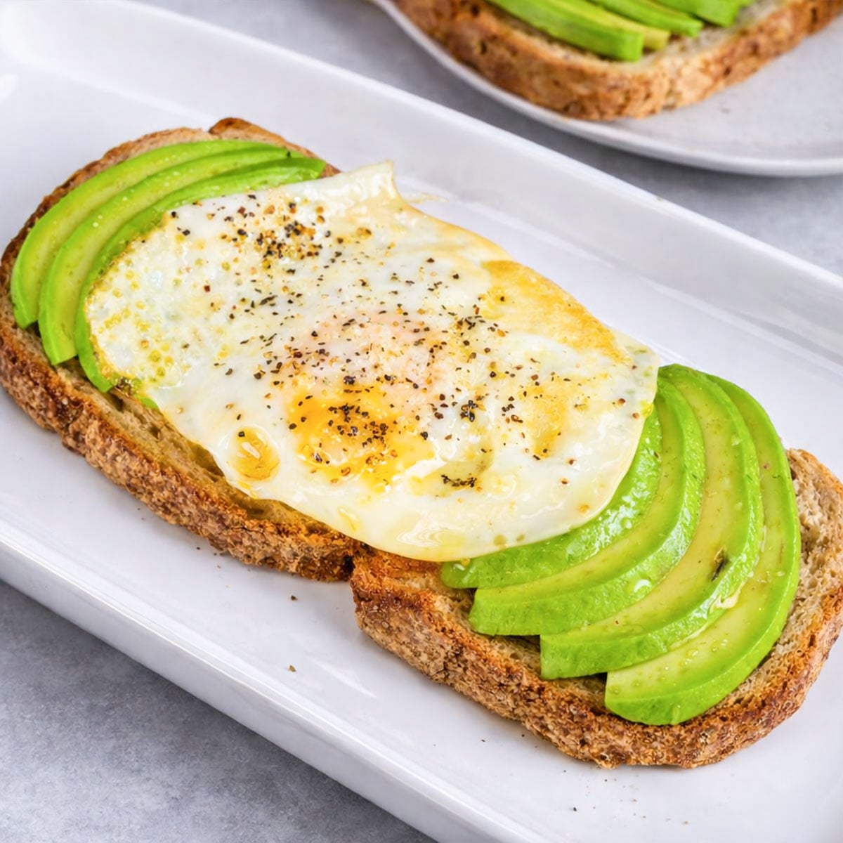 Avocado toast with over-easy egg on sourdough, topped with black pepper on white plate