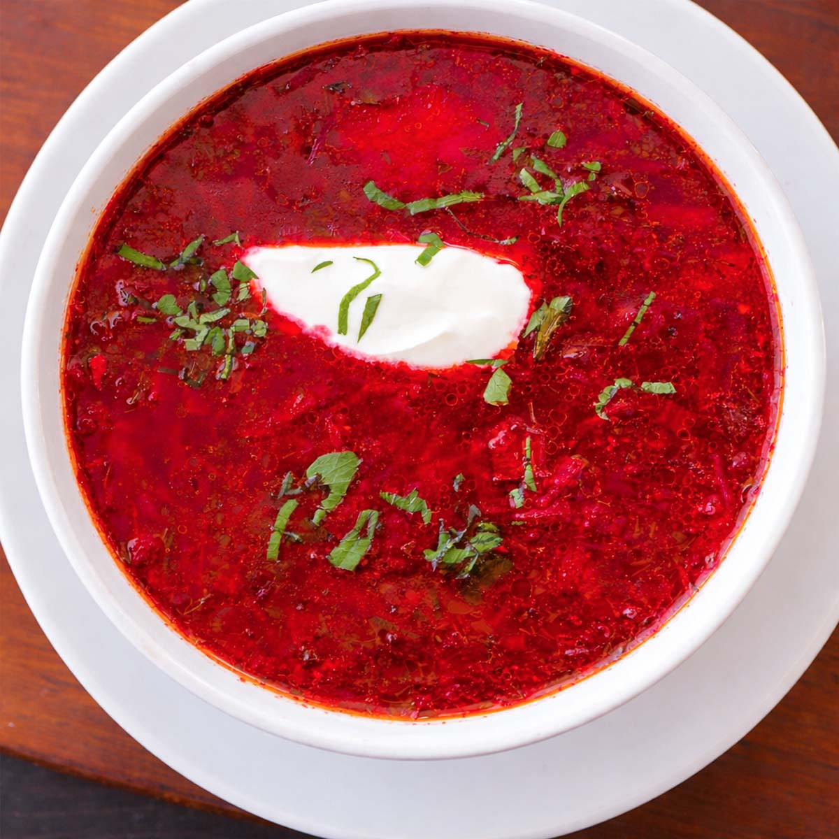Overhead view of Ukrainian borscht beet soup in a white bowl, topped with sour cream and fresh herbs.