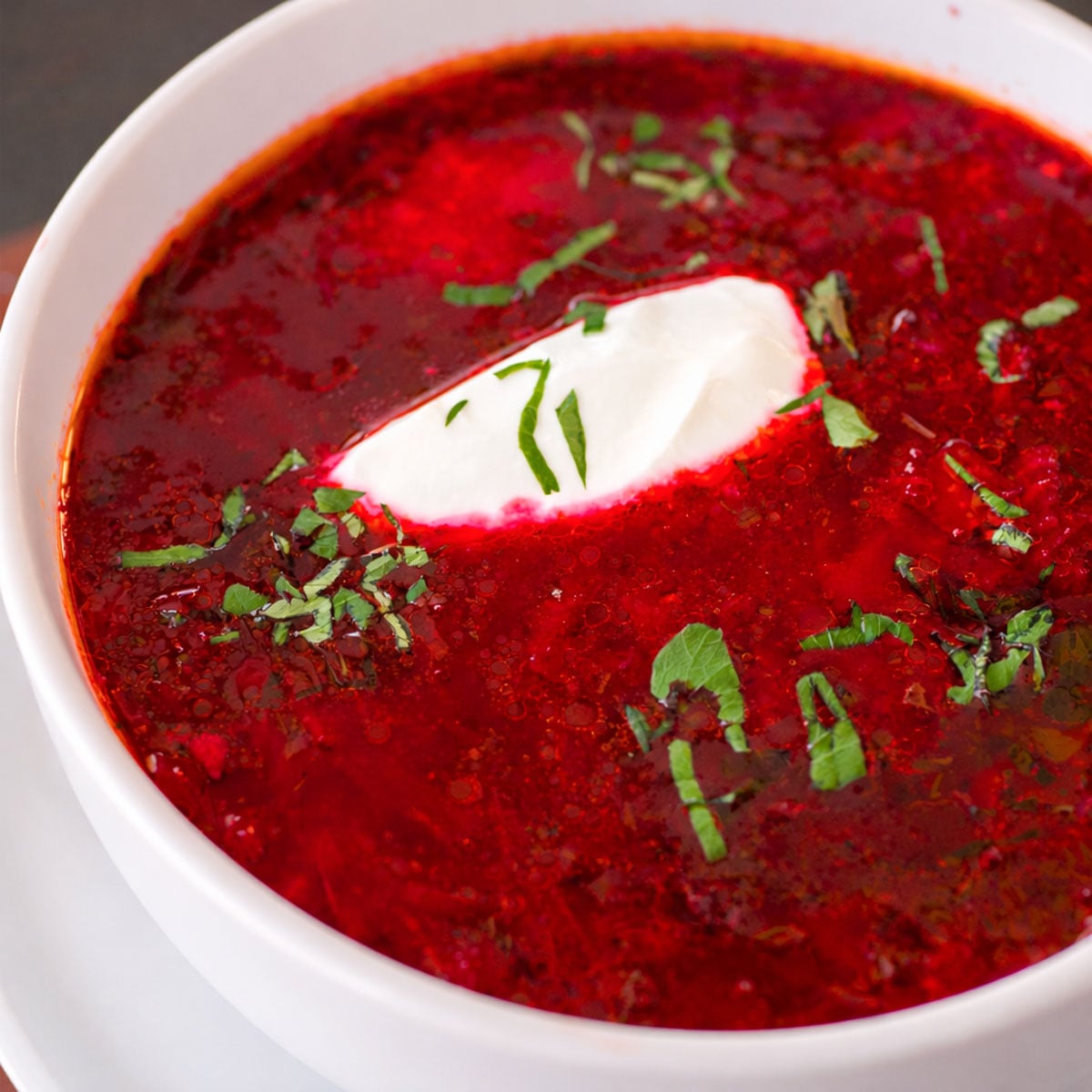 Close-up of borscht beet soup in a white bowl, topped with a dollop of sour cream and chopped fresh herbs.