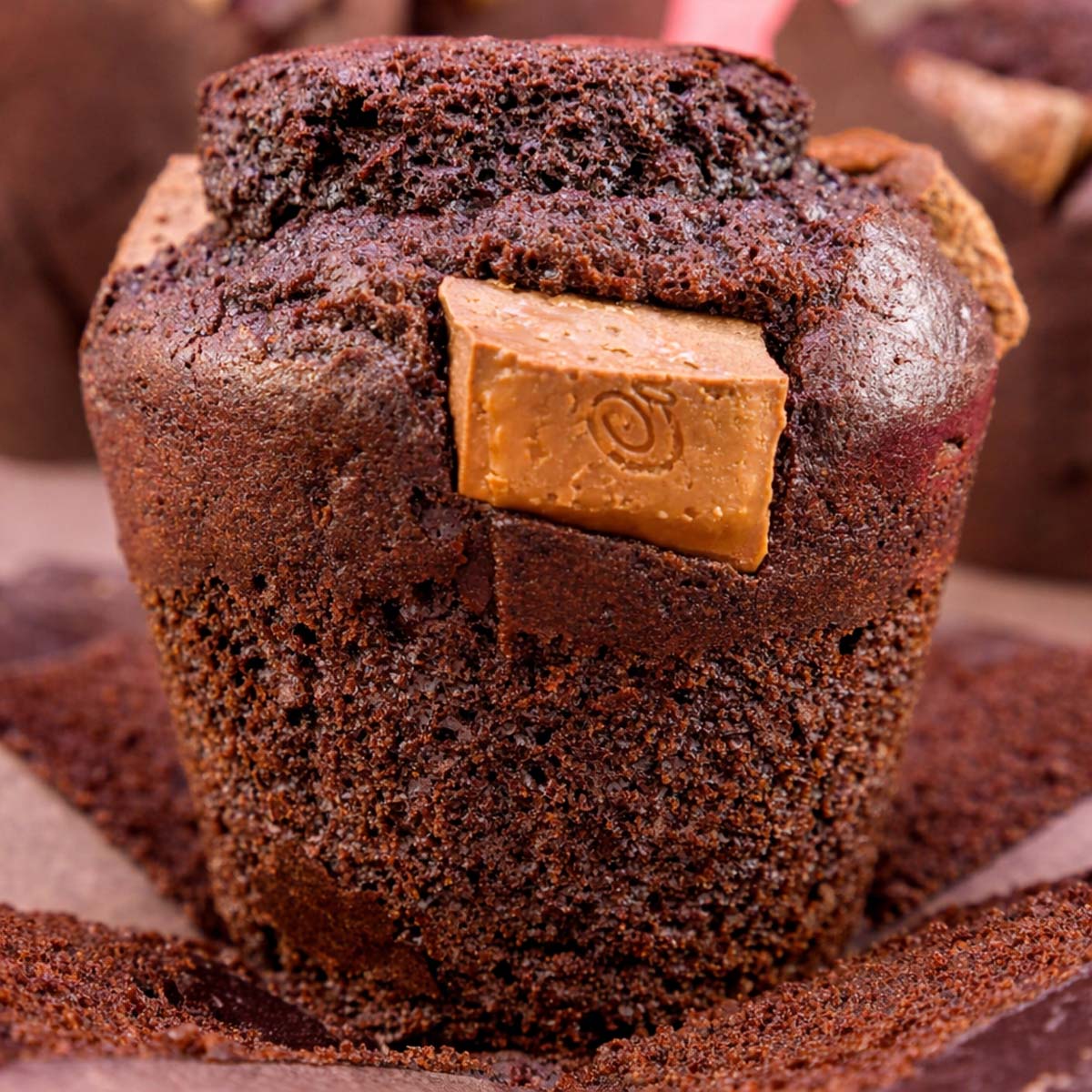 Close-up of a rich chocolate muffin with a milk chocolate square baked into the top, sitting in a brown paper liner.