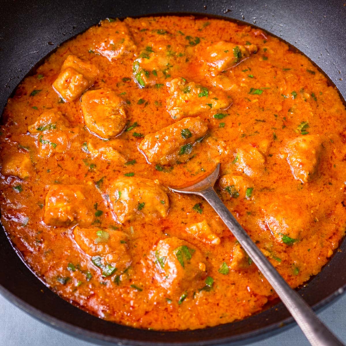 Creamy coconut chicken curry simmering in a black pan with a spoon, finished with chopped cilantro