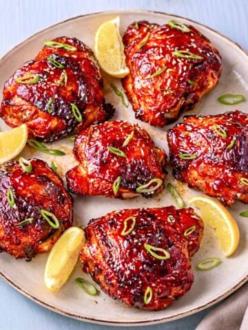 Gochujang chicken thighs on a plate with sesame seeds, sliced green onions, and lemon wedges