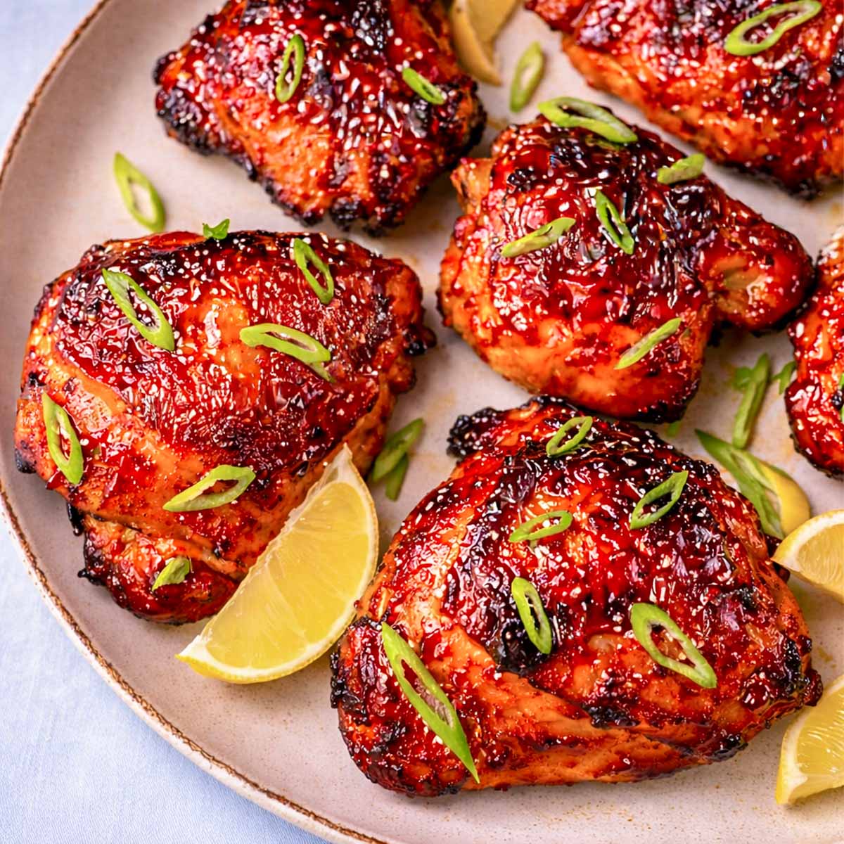 Close-up of gochujang chicken thighs with, sliced green onions, and lemon wedges on a ceramic plate