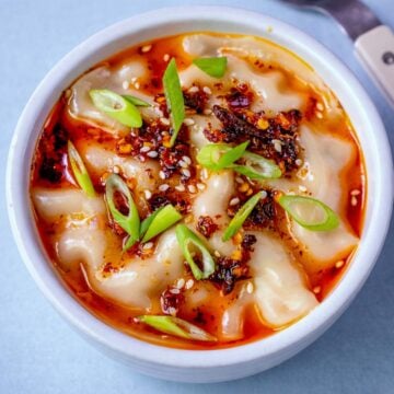 Pork dumpling lasagna topped with chili oil, sesame seeds, and sliced green onions in a white bowl.