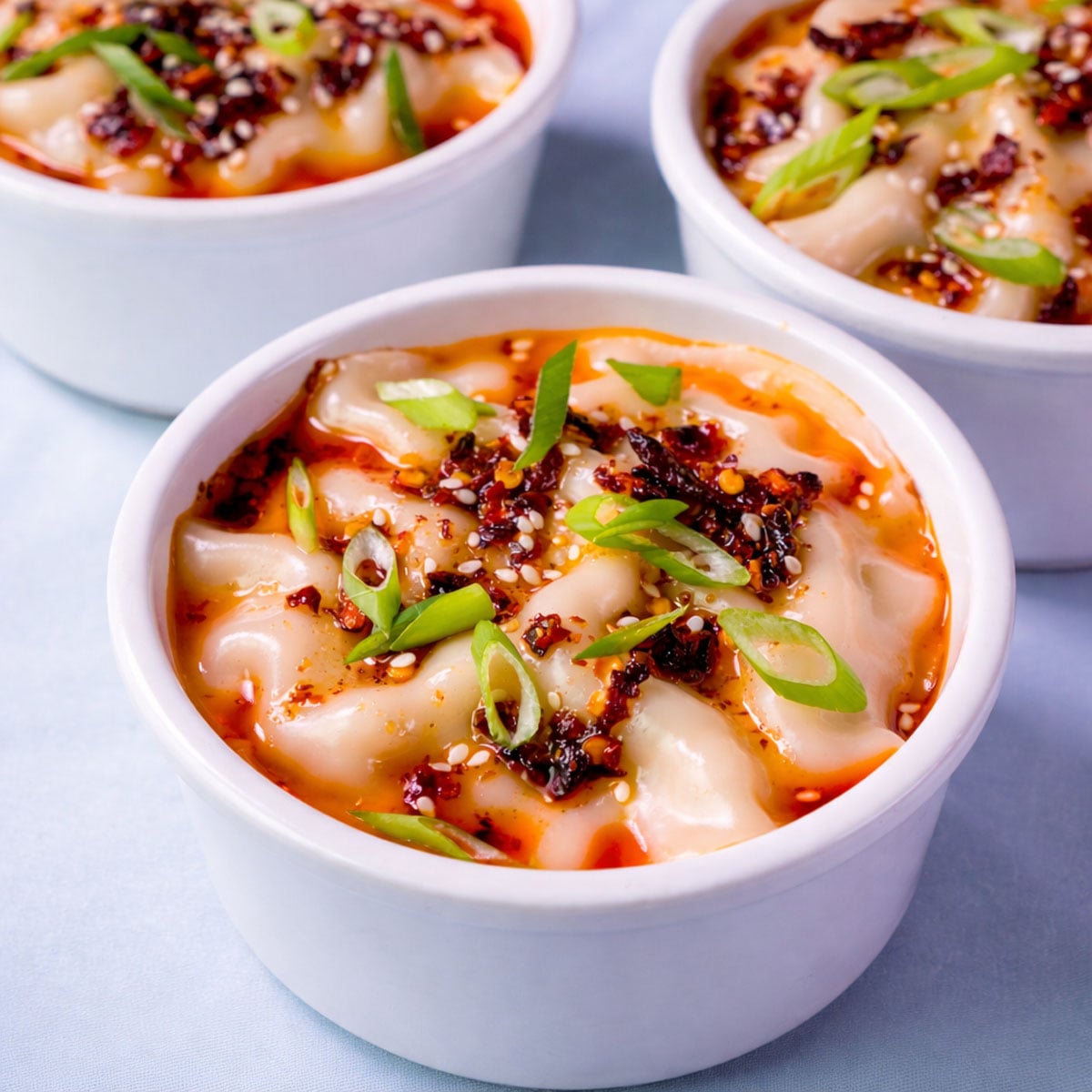Three bowls of pork dumpling lasagna topped with chili oil, sesame seeds, and sliced green onions.