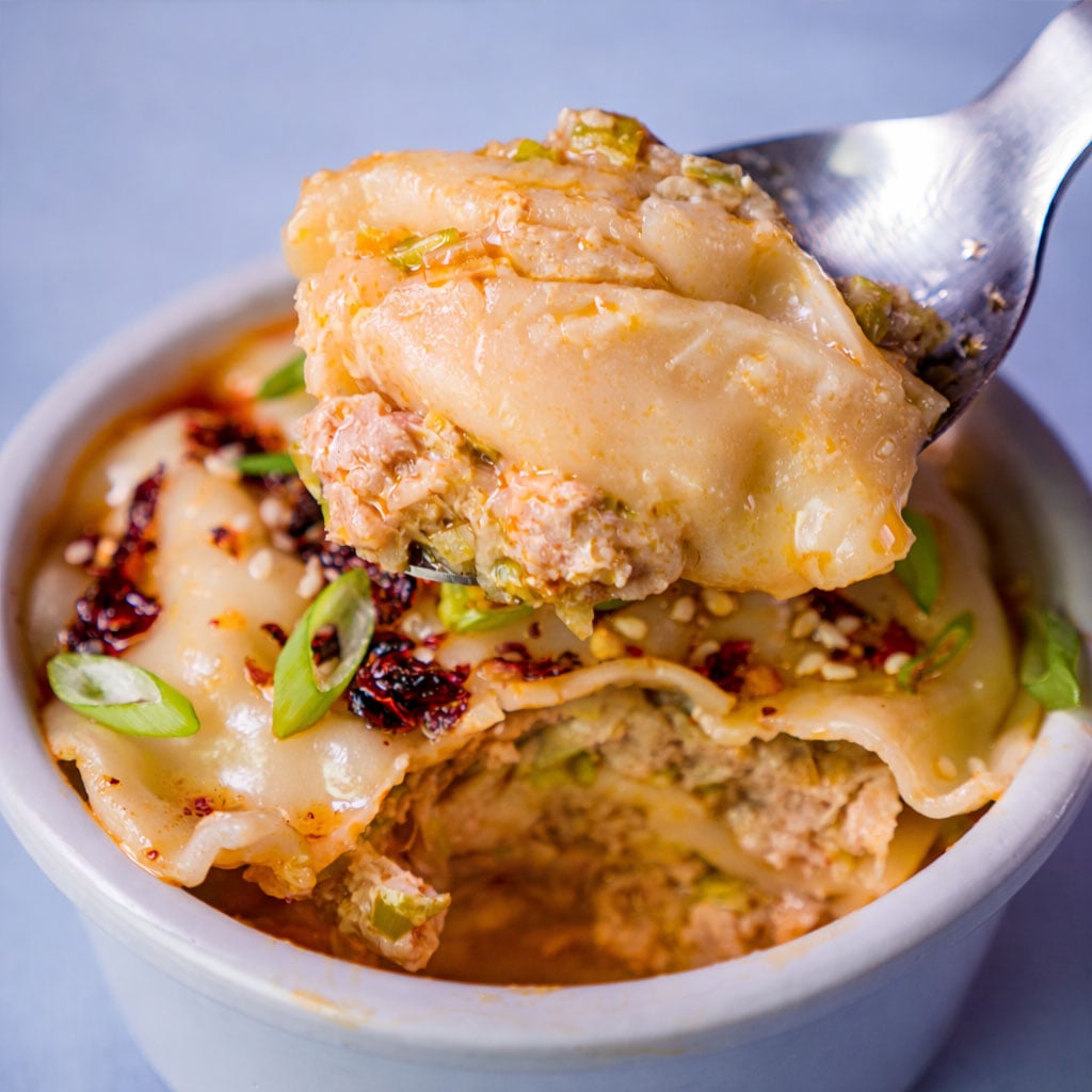 Spoonful of pork dumpling lasagna lifted from a bowl, showing the savory filling, with sesame seeds, chili oil, and green onions on top.