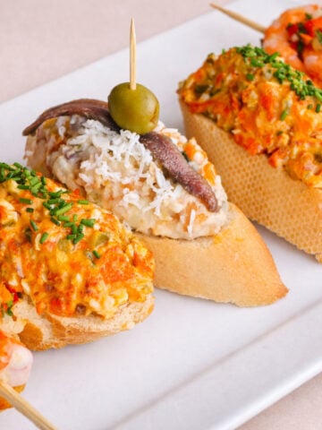 3 Spanish pintxos (pinchos) on a white platter: a shrimp-topped baguette with chopped peppers, an anchovy-and-olive skewer over a creamy potato salad topping, and a baguette topped with a chorizo-style salad and chives.