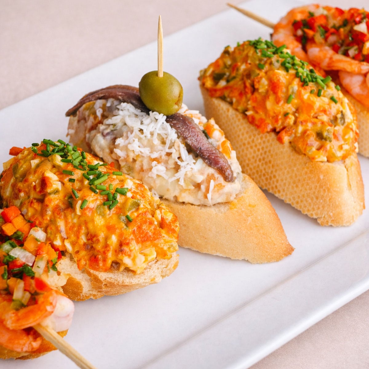 3 Spanish pintxos (pinchos) on a white platter: a shrimp-topped baguette with chopped peppers, an anchovy-and-olive skewer over a creamy potato salad topping, and a baguette topped with a chorizo-style salad and chives. 