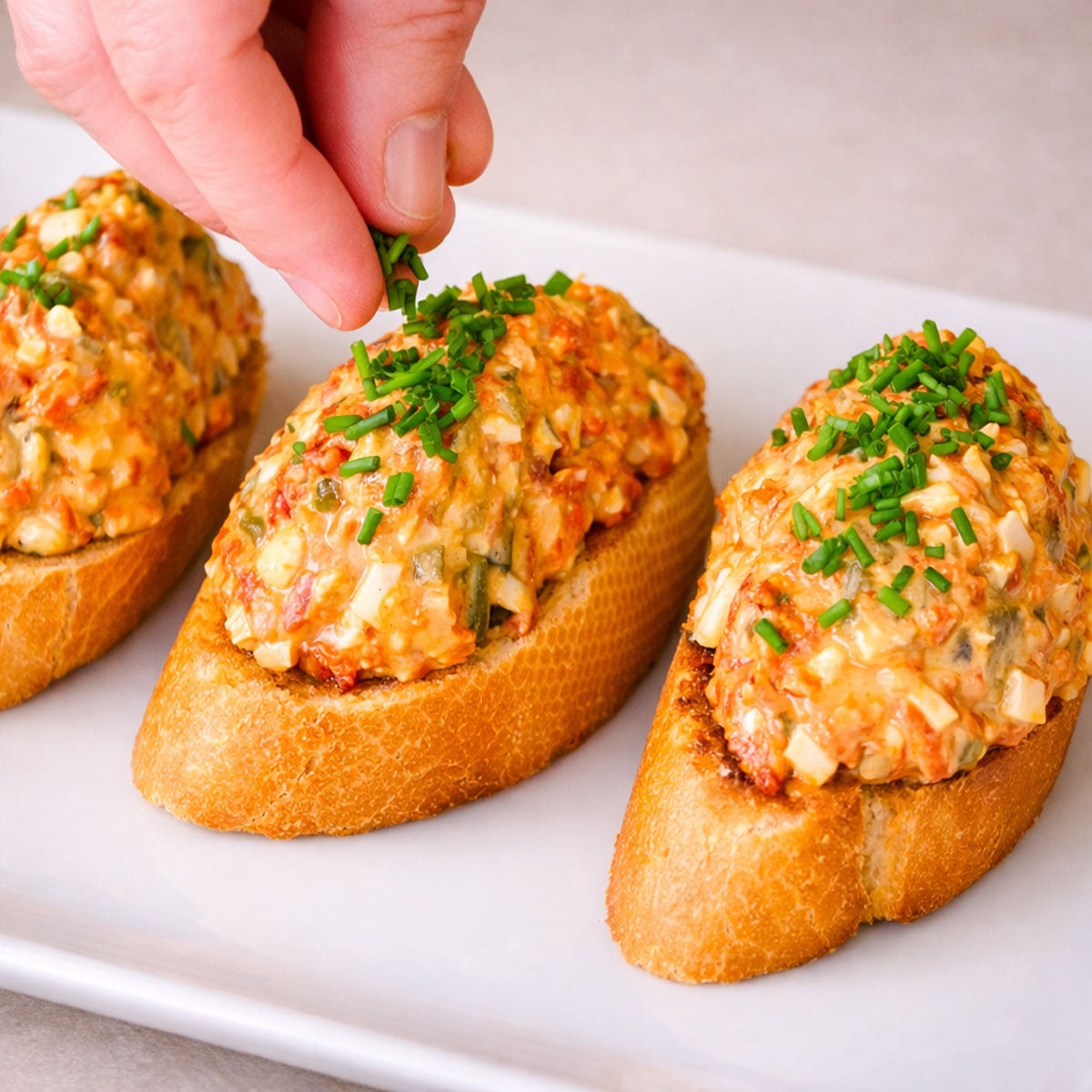 Hand sprinkling chopped chives over chorizo pintxos, with creamy chorizo salad spooned onto toasted baguette slices. 