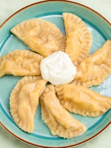 Apple pierogi arranged in a circle on a teal plate with a dollop of sour cream in the center on an orange tabletop.