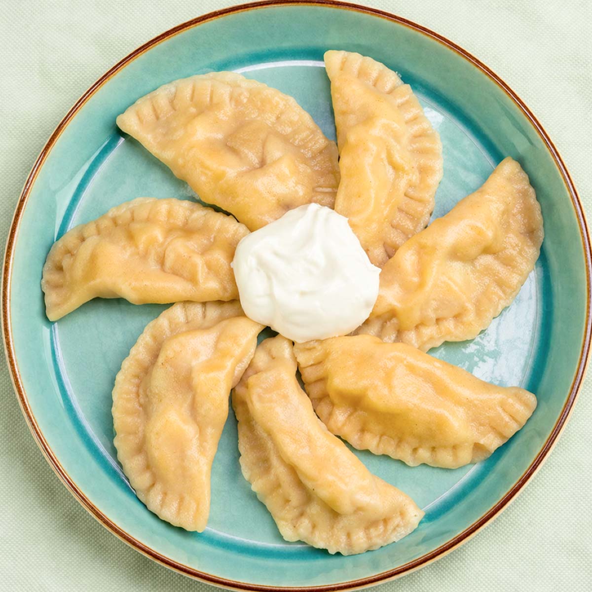 Apple pierogi arranged in a circle on a teal plate with a dollop of sour cream in the center.