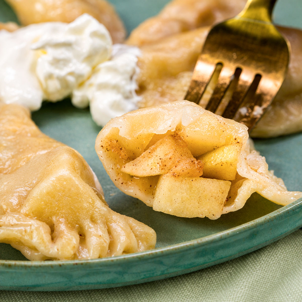 Close-up of an apple pierogi opened to show warm cinnamon apple filling, held on a fork beside a dollop of sour cream.
