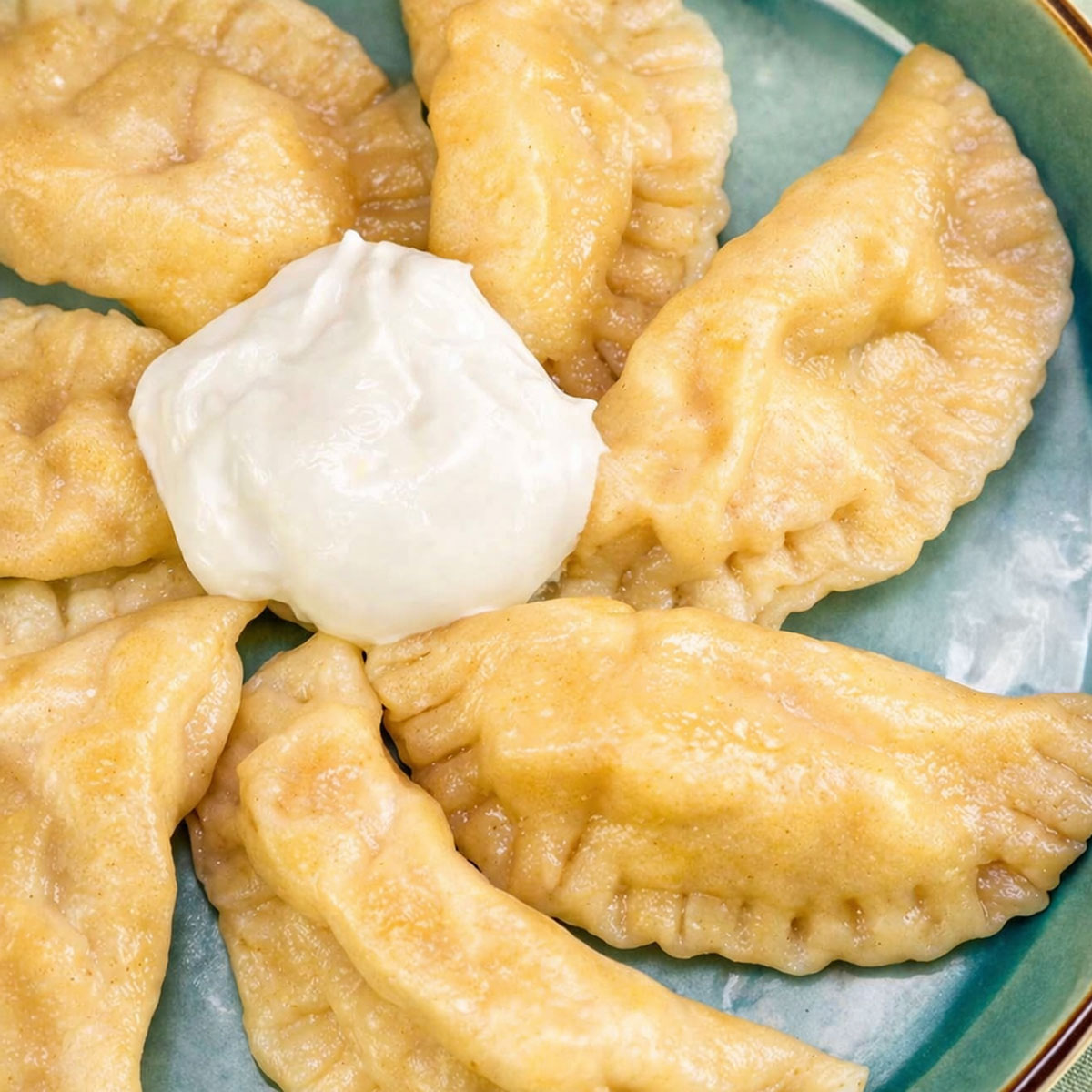 Apple pierogi arranged on a teal plate with a dollop of sour cream in the center. 