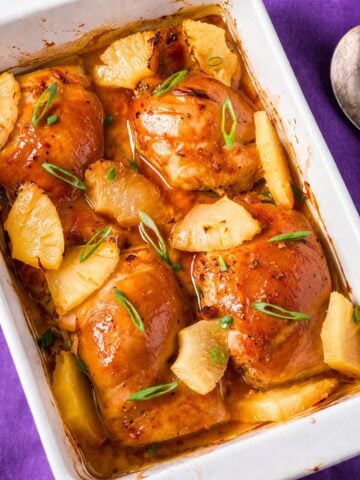 Baked pineapple chicken thighs in a white dish with caramelized glaze, juicy pineapple chunks, and fresh green onion garnish on a purple background