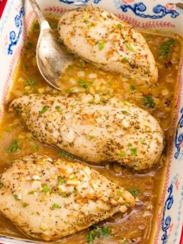 Baked chicken in chicken stock in a ceramic dish with garlic, herbs, and onions, served in a rich golden sauce with a spoon resting on the side