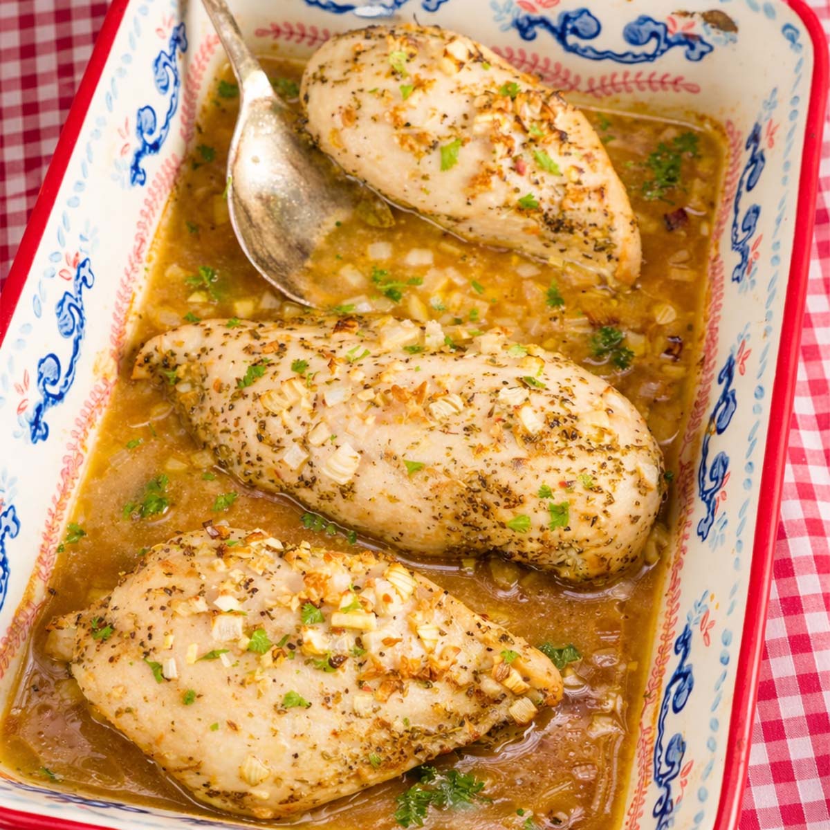 Baked chicken in chicken broth in a ceramic dish with garlic, herbs, and onions, served in a rich golden sauce with a spoon resting on the side