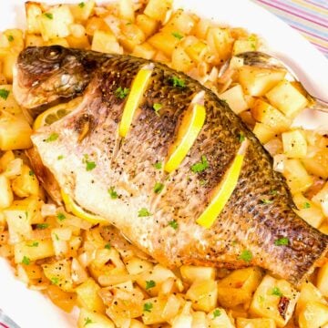 Baked whole tilapia with lemon slices on top, served on a white platter over roasted potatoes and onions