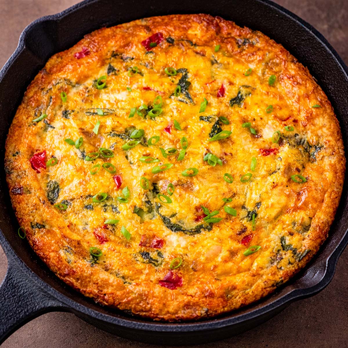 Chicken frittata baked in a cast iron skillet with spinach, red peppers, melted cheese, and fresh green onions on top