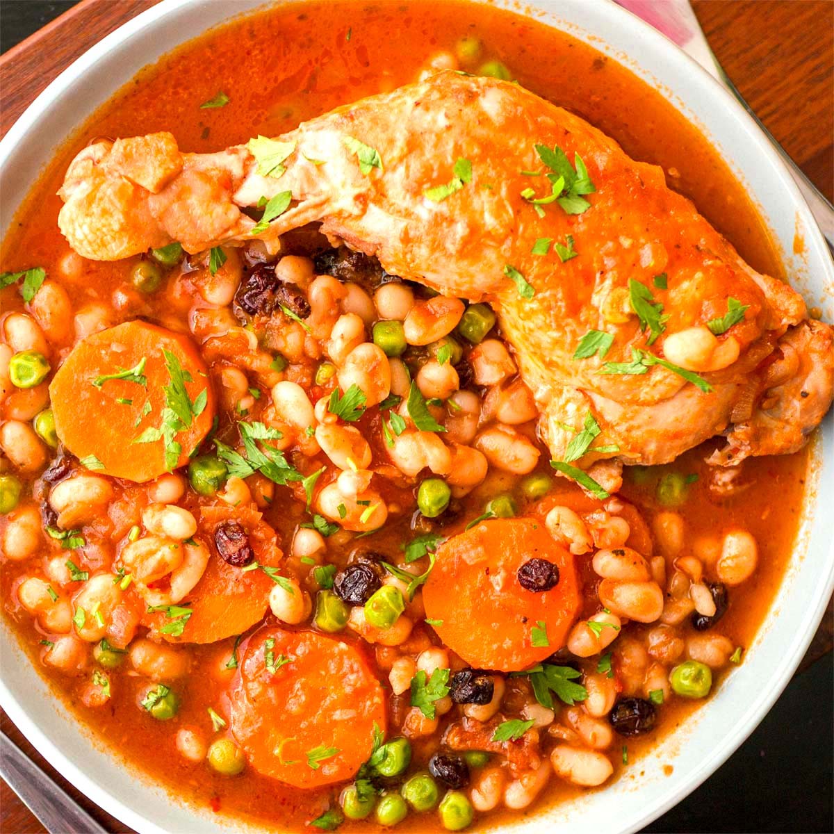 Chicken and bean stew with carrots, peas, and tomato sauce served with a tender chicken leg in a bowl, garnished with fresh parsley.