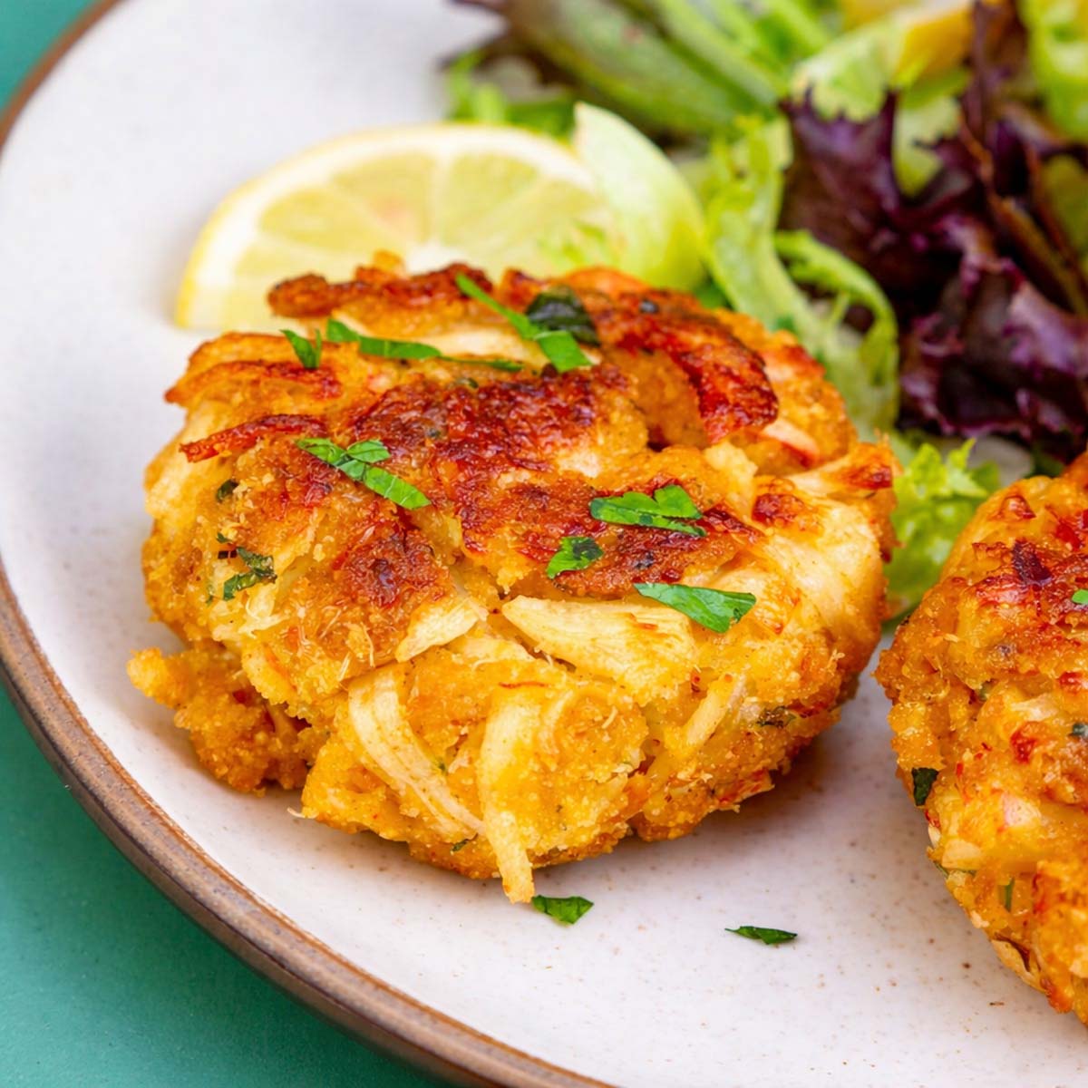 Crispy canned crab cakes on a white plate with chopped parsley, a lemon wedge, and mixed greens on the side. 