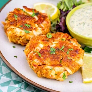 Two golden crab cakes on a plate with chopped parsley, lemon wedges, mixed greens, and a bowl of creamy dipping sauce.