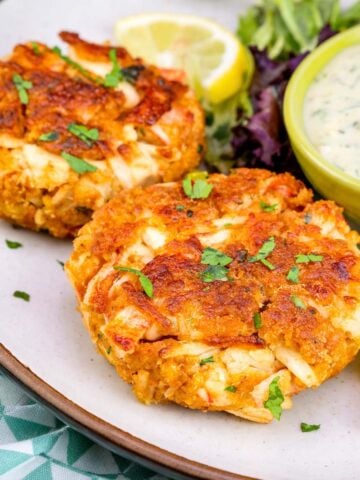 Two golden crab cakes on a plate with chopped parsley, lemon wedges, mixed greens, and a bowl of creamy dipping sauce.